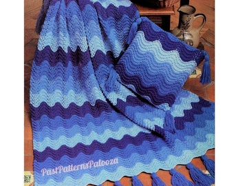 May include: A crocheted blanket and cushion set in shades of blue with a wavy pattern. The blanket and cushion have blue tassels along the edges. The set is displayed on a tiled floor.