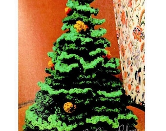 Op de afbeelding: Een groene gehaakte kerstboom met gele en oranje ornamenten. De boom staat op een bruine tafel met een bloemige achtergrond.