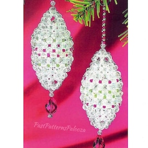 以下が含まれることがあります： ピンクとグリーンのアクセントが付いた白いビーズでできたクリスマスオーナメントが2つ。オーナメントはクリスマスツリーの枝にぶら下がっています。オーナメントの下には、涙滴型のクリスタルビーズが付いています。