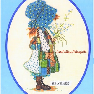 May include: Cross stitch pattern featuring a girl in a blue and white dress holding a bouquet of flowers. The pattern is titled "Holly Hobbie" and is from PastPatternPaloozaCo.