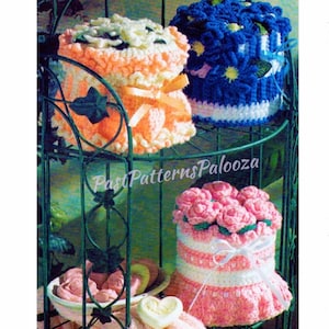 May include: Three crocheted toilet paper roll covers in a wire rack. The top cover is blue with white flowers, the middle cover is peach with white flowers, and the bottom cover is pink with white flowers.  PastPatternsPalooza is printed on the rack.