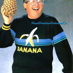May include: A man wearing a black sweater with a yellow banana graphic and the word "BANANA" in yellow letters. He is holding a pineapple in his left hand and smiling.
