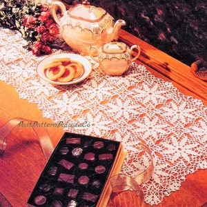Puede incluir: Un camino de mesa de crochet blanco con un diseño floral. El camino de mesa está colocado sobre una mesa de madera con una tetera, un azucarero, galletas y una caja de chocolates.