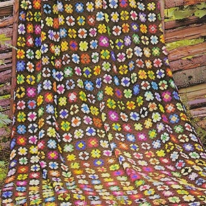 May include: A colorful crochet blanket with a brown background and multi-colored flowers. The blanket is made with granny squares and is draped over a wooden fence.