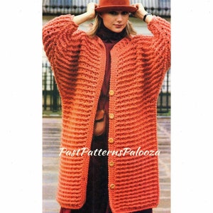 May include: A long, orange crocheted cardigan with a button closure. The cardigan has a ribbed texture and is worn by a model with a red hat.