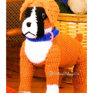 May include: A crocheted stuffed boxer dog toy with brown, white, and black fur. The dog is wearing a blue and pink collar. The dog is standing on a wooden surface.