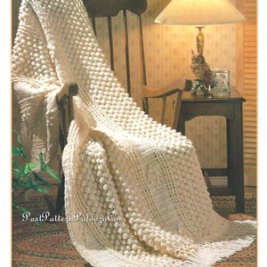 May include: A cream-coloured, textured blanket draped over a wooden rocking chair. The blanket has a bobble stitch design and a fringed edge. A lamp with a beige shade and an owl figurine are on a side table. The text "Past Pattern Palooza Co" is visible.