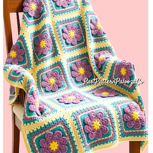 Op de afbeelding: Een handgemaakte gehaakte deken, gedrapeerd over een houten stoel. De deken heeft een herhalend bloemenpatroon met paarse, turquoise, gele en crèmekleurige vierkanten. De tekst "PastPatternPaloozaCo" is zichtbaar in de rechterbovenhoek.