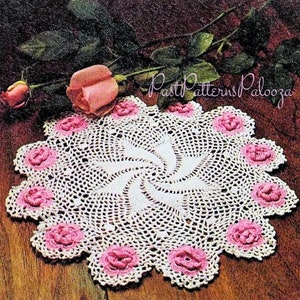 Könnte beinhalten: Weiße Häkeldeckchen mit einem rosafarbenen Rosenmuster. Die Deckchen sind rund und haben ein Spiralmuster in der Mitte. Die Deckchen haben einen Durchmesser von etwa 35 cm.