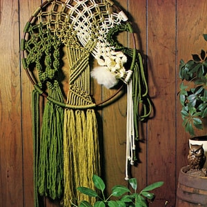 Op de afbeelding: Een macramé wandhanger met een ronde houten frame, met een groen en wit ontwerp met lange franjes. De hanger is bevestigd aan een houten paneelwand.