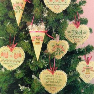 K&ouml;nnte beinhalten: Eine Sammlung von cremefarbenen Kreuzstich-Anh&auml;ngern in Herz- und Kegelform mit roten und gr&uuml;nen Weihnachtsmotiven. Ein herzf&ouml;rmiger Anh&auml;nger zeigt den Text 'We wish you a Merry Christmas and a Happy New Year' und ein anderer 'Noel'.