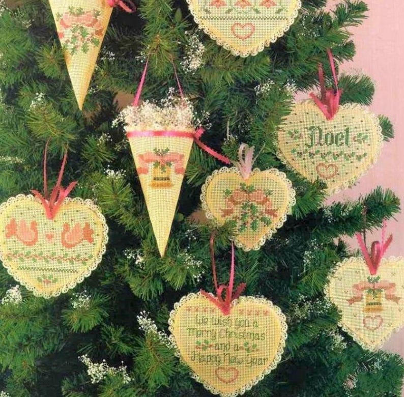 K&ouml;nnte beinhalten: Eine Sammlung von cremefarbenen Kreuzstich-Anh&auml;ngern in Herz- und Kegelform mit roten und gr&uuml;nen Weihnachtsmotiven. Ein herzf&ouml;rmiger Anh&auml;nger zeigt den Text 'We wish you a Merry Christmas and a Happy New Year' und ein anderer 'Noel'.