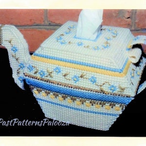 以下が含まれることがあります： 青と黄色のアクセントが付いた、白い編み物でできたティーポット型のティッシュボックスカバー。カバーには花柄のデザインと、上部にティッシュ用の開口部があります。