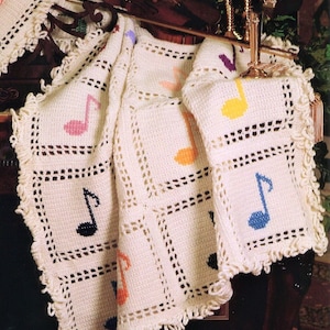 Può includere: Coperta all'uncinetto color crema con note musicali colorate. La coperta ha un motivo quadrato e un bordo decorativo. Le note musicali sono in vari colori, tra cui rosa, arancione, giallo e blu. La coperta è drappeggiata su una superficie scura.