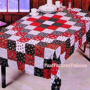 May include: A red, white, and black patchwork tablecloth with a floral pattern. The tablecloth is laid out on a wooden table with a chair on either side. The tablecloth is made of fabric squares sewn together.