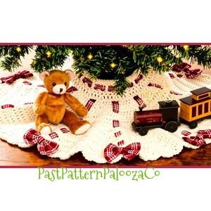 May include: A round, cream-colored crocheted tree skirt featuring a scalloped edge and red and white plaid fabric bows woven throughout. A brown plush teddy bear and a small wooden toy train are positioned on top of the fabric surface.