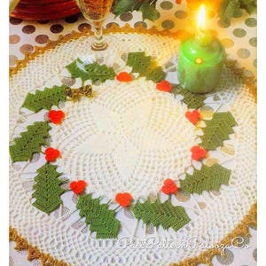 Pode incluir: Um crochê branco com uma borda dourada e folhas de azevinho verdes com bagas vermelhas ao redor da borda. O crochê está sobre uma toalha de mesa branca com bolinhas pretas.