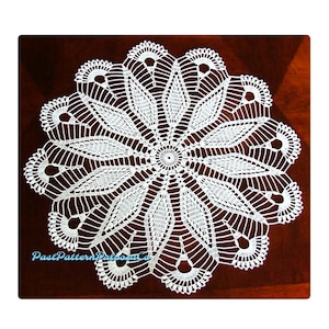 Puede incluir: Doily de ganchillo blanco con un delicado diseño floral. El doily es circular y tiene un centro con un círculo más pequeño. El doily está sobre una superficie marrón oscura.
