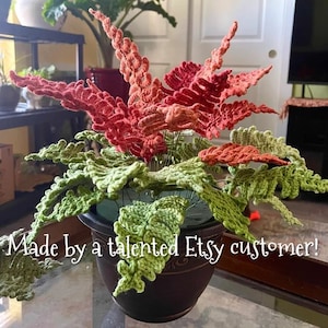 K&ouml;nnte beinhalten: Eine geh&auml;kelte Farnpflanze in einem braunen Topf mit gr&uuml;nen und orangen Bl&auml;ttern. Die Pflanze steht auf einem Glastisch. "Made by a talented Etsy customer!" steht auf dem Tisch.