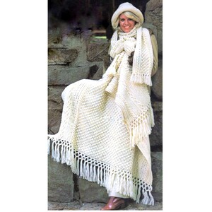 May include: A white crocheted blanket with fringe. The blanket is draped over a person wearing a white hat and scarf.