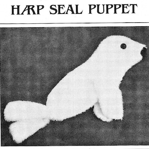 May include: A white harp seal puppet with black eyes. The puppet is made of soft, plush fabric and has a long, slender body with flippers.