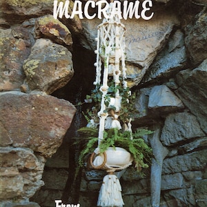 May include: A white macrame plant hanger with a white pot and green plant hanging from a stone wall. The text "A Sip Of Macrame' From Brandy's" is printed on the image.