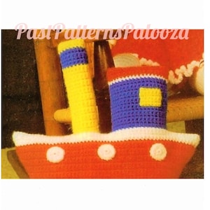 Pode incluir: Um barco de brinquedo de crochê com um casco vermelho, guarnições brancas e um mastro listrado azul e branco. O barco tem três círculos brancos no casco e um quadrado amarelo no mastro azul.