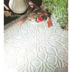 May include: A white, intricately crocheted doily with a circular floral pattern. The doily is displayed on a table with a white pitcher, a small bowl of candies, and a pink ribbon. The text "Past Pattern Palooza Co" is visible.