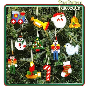 May include: A collection of twelve plastic canvas Christmas ornaments featuring a train, trumpet, Santa face, bell, snowman, tree, two nutcrackers, candle, candy cane, dove, and stocking, all with white hanging strings and PastPatternPaloozaCo text.
