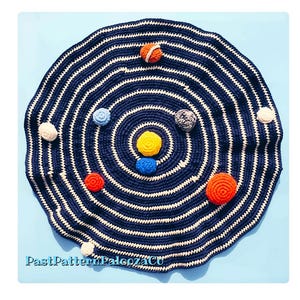 Peut inclure: Un tapis rond en crochet bleu foncé et blanc avec un motif en spirale et plusieurs petites planètes sphériques en crochet colorées fixées sur la surface. Le texte "PastPatternPaloozaCo" est imprimé en bleu clair en bas.