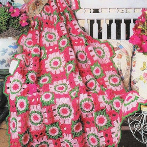 May include: A crocheted blanket with pink and green squares, featuring a floral design. The blanket is draped over a white bench with a floral patterned pillow.