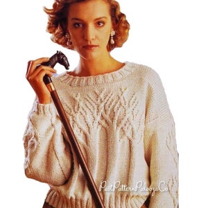 Może przedstawiać: Kremowy sweter z dzianiny z wzorem przypominającym drzewo z przodu. Sweter ma okrągły dekolt oraz prążkowane mankiety i dół. W dłoni trzymana jest ciemnobrązowa laska z uchwytem w kształcie głowy konia.