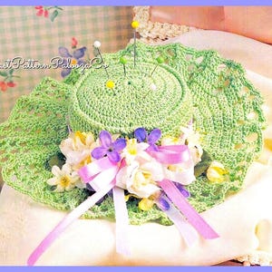 Puede incluir: Un sombrero de ganchillo verde con un borde de encaje blanco y flores moradas y blancas. El sombrero se utiliza como alfiletero y tiene varios alfileres que sobresalen de él.