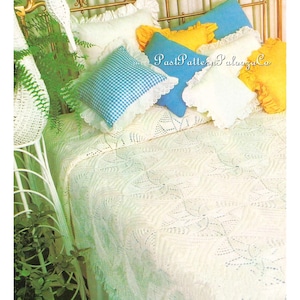 May include: A bed with a cream-colored, open-weave bedspread and matching pillows. The pillows are in various colors, including blue, yellow, and white, with lace trim. A white wicker plant stand with greenery is on the left.