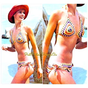 May include: A crochet bikini set with a colourful geometric pattern. The top is a halter style with a triangle shape in the front. The bottom is a tie-side style with a similar geometric pattern. The model is wearing a red hat.