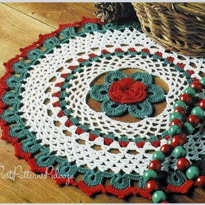 May include: A white crochet doily with a green and red border and a red crocheted flower in the center. The doily is on a wooden surface with a basket and beads in the background.