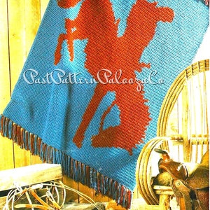 May include: A crocheted blanket with a blue background and a red silhouette of a person riding a horse. The blanket has fringe around the edges. The blanket is draped over a wooden chair with a saddle on it.