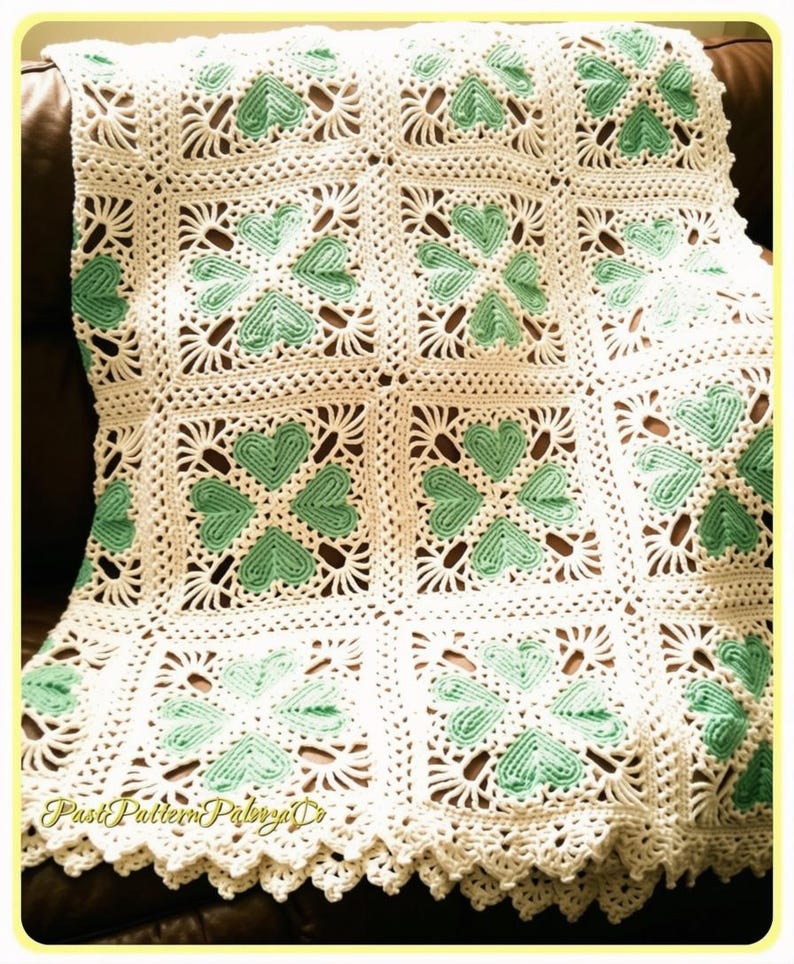 May include: A handmade crochet blanket featuring a repeating square motif pattern. Each square contains four mint green heart-shaped elements arranged in a clover design, surrounded by white yarn. The blanket has a decorative scalloped edge.