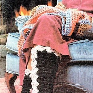 May include: A woman wearing black and white crocheted slippers with a pink trim sits in a blue armchair by a fireplace. She is wearing a pink robe and crocheting a multi-colored blanket.