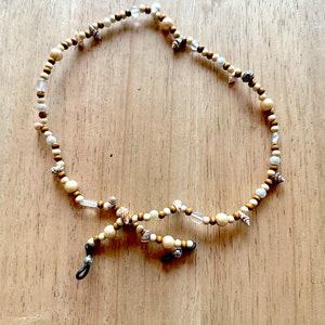 May include: Eyeglass chain with a mix of wooden beads, small seashells, and clear glass beads. The chain is a light beige color with dark brown and clear accents. The chain is designed to hold eyeglasses.