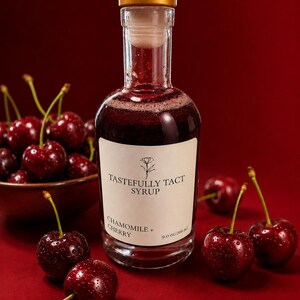 Puede incluir: Una botella de vidrio transparente con jarabe rojo oscuro, tapa dorada y etiqueta blanca que dice TASTEFULLY TACT SYRUP CHAMOMILE + CHERRY 9.0 oz/266 ml. La botella está rodeada de cerezas frescas de color rojo oscuro con tallos verdes.