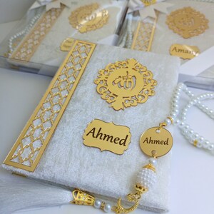 May include: A close-up of a white and gold religious book with the name "Ahmed" on a gold plaque. The book has a gold filigree design and is accompanied by a pearl beaded prayer beads. Other similar books are visible in the background.