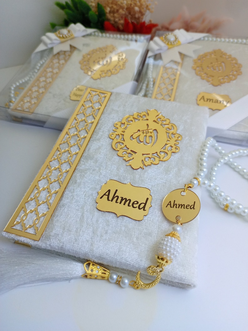 May include: A close-up of a white and gold religious book with the name "Ahmed" on a gold plaque. The book has a gold filigree design and is accompanied by a pearl beaded prayer beads. Other similar books are visible in the background.