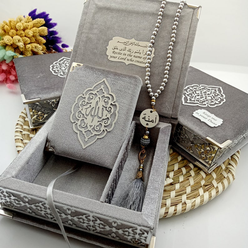 May include: A gray velvet box with intricate silver designs, containing a small gray book with a silver Arabic calligraphy design, a gray prayer bead necklace with a silver charm, and a gray tassel. The box has a silver inscription that reads "Recite in the name of your Lord who created" in Arabic and English.