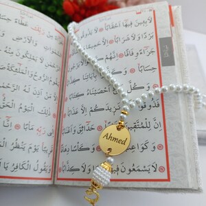 May include: A pearl prayer bead with a gold pendant engraved with the name "Ahmed" hangs over an open book. The book's pages display Arabic script. A tassel and decorative elements complete the prayer bead.