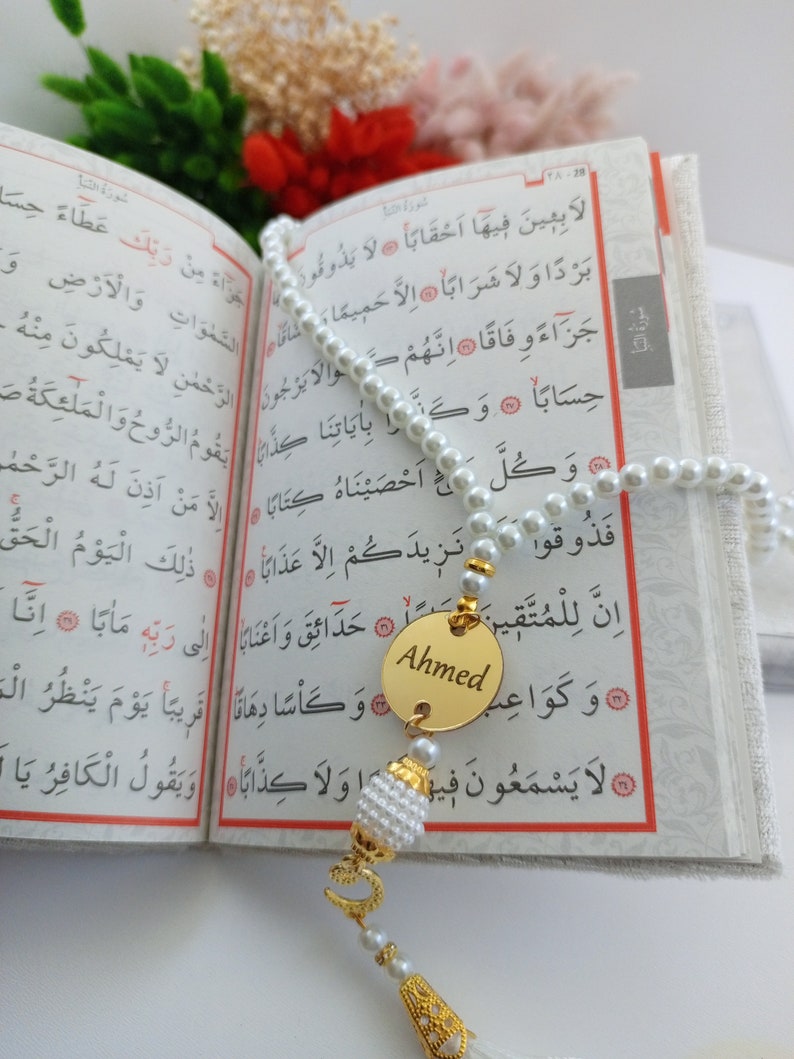 May include: A pearl prayer bead with a gold pendant engraved with the name "Ahmed" hangs over an open book. The book's pages display Arabic script. A tassel and decorative elements complete the prayer bead.