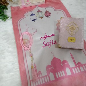 May include: A pink prayer rug with a white center featuring a mosque, lanterns, and the name "Safia" in Arabic script. The rug is decorated with a pink tassel and a pink prayer bead necklace. A pink velvet prayer book with the name "Safia" on the cover is also included.