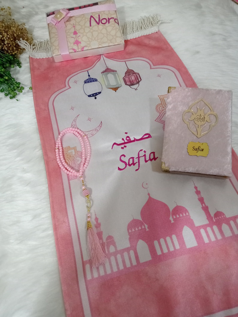 May include: A pink prayer rug with a white center featuring a mosque, lanterns, and the name "Safia" in Arabic script. The rug is decorated with a pink tassel and a pink prayer bead necklace. A pink velvet prayer book with the name "Safia" on the cover is also included.