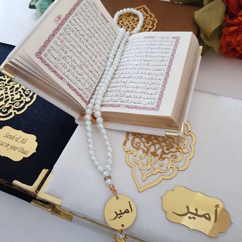 May include: A white prayer rug with gold accents and a gold decorative design. A white prayer bead necklace with a gold pendant is on the rug. The rug has the words "Sarah & Ali" and "us in your Duas" written on it. The rug is folded in half and a small book is on top of it.