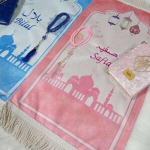 May include: Two prayer rugs with white fringe, one blue and one pink. The blue rug has the name "Bilal" written on it and the pink rug has the name "Safia" written on it. Both rugs feature a mosque design and a crescent moon.